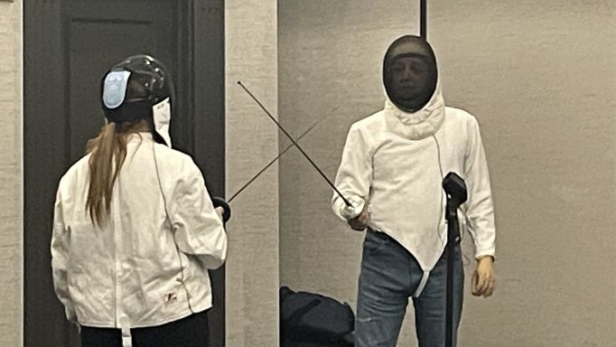 A photo of two people fencing.  They are facing each other, each wearing a protective jacket and head shield.  Each is holding a sword.  The swords are pointed slightly upwards and are crossed in preparation for starting a round.